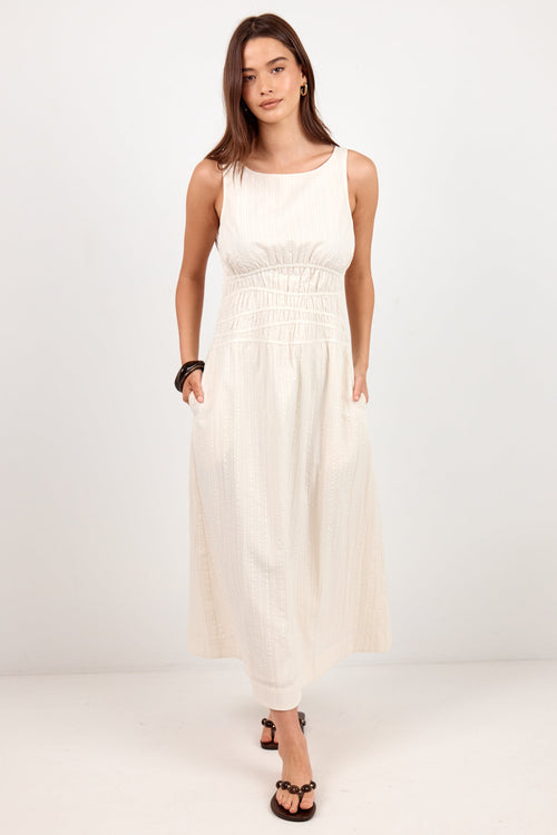 A woman modeling a sleeveless, off-white midi dress with a smocked bodice and side pockets, paired with black sandals, against a plain white studio background.