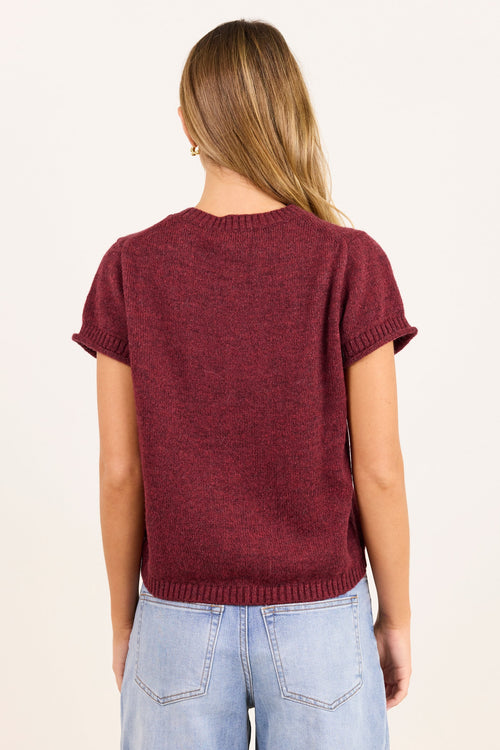 A woman viewed from behind wearing a short-sleeve maroon knit top and light blue jeans against a plain off-white background.
