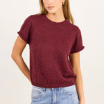 A woman modeling a short-sleeve crewneck knit sweater in heathered burgundy, paired with light-wash blue jeans, against a plain off-white studio background.