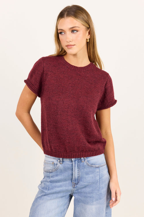 A woman modeling a short-sleeve crewneck knit sweater in heathered burgundy, paired with light-wash blue jeans, against a plain off-white studio background.