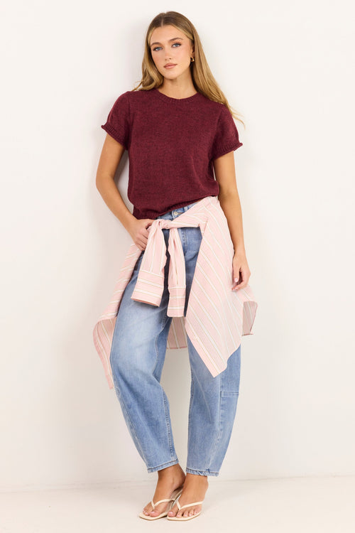 A woman modeling a dark burgundy short-sleeve t-shirt, light-wash blue jeans, and a light pink striped shirt tied around her waist, standing against a plain white studio background.