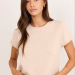 A woman modeling a short-sleeve crewneck knit top in a light beige color, paired with blue jeans and a brown belt, against a plain light background.