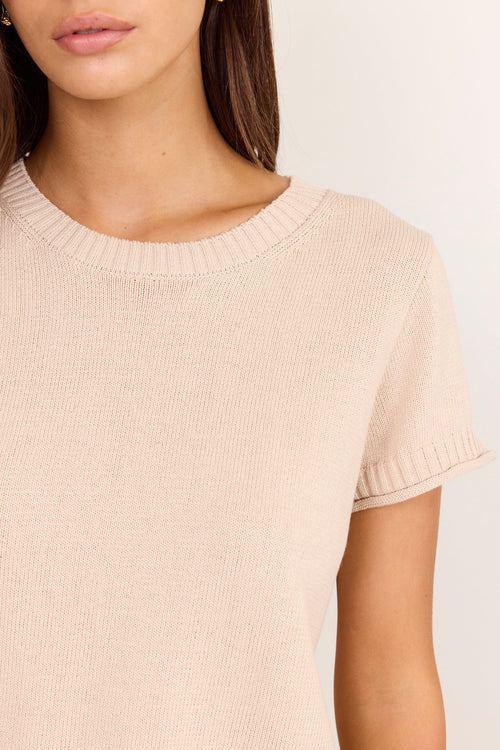 Close-up of a woman wearing a light beige short-sleeve knit top with a crew neck and ribbed sleeve cuffs, against a plain light background.