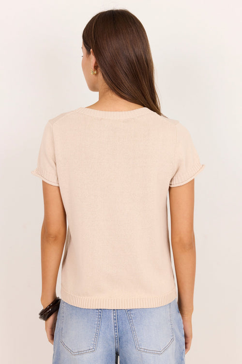A woman viewed from the back wearing a light beige short-sleeve knit top and light blue jeans against a plain off-white background.