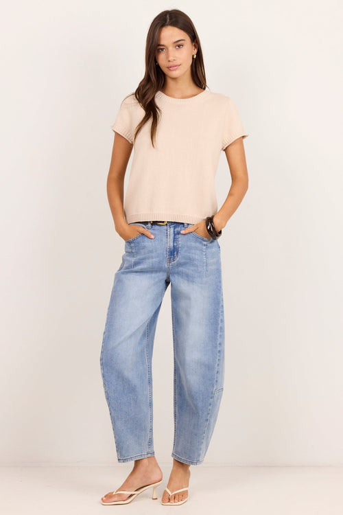 A woman modeling a light beige short-sleeve crop top and light-wash blue wide-leg jeans, standing against a plain off-white studio background.