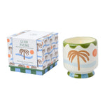 A Lush Palms scented soy wax candle in a ceramic cup with a tropical palm tree design, shown next to its patterned retail box on a white background.