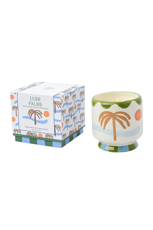 A Lush Palms scented soy wax candle in a ceramic cup with a tropical palm tree design, shown next to its patterned retail box on a white background.
