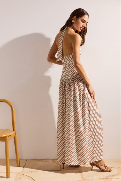 Woman in a striped dress standing next to a wooden chair against a plain wall.