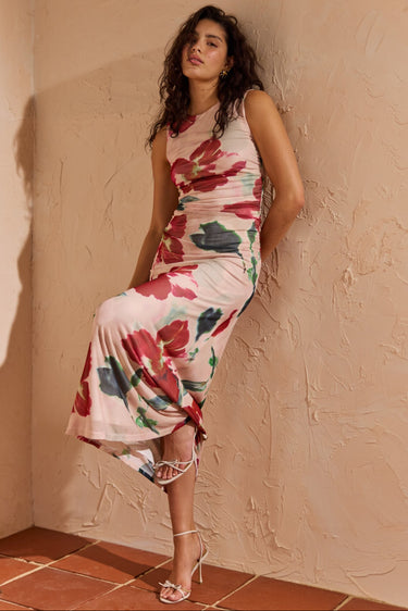 Woman in a floral dress standing against a textured wall with a small table beside her.