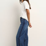 Woman wearing mid-blue wide-leg jeans with a dark side stripe and white t-shirt, styled with white sneakers against a neutral wall.