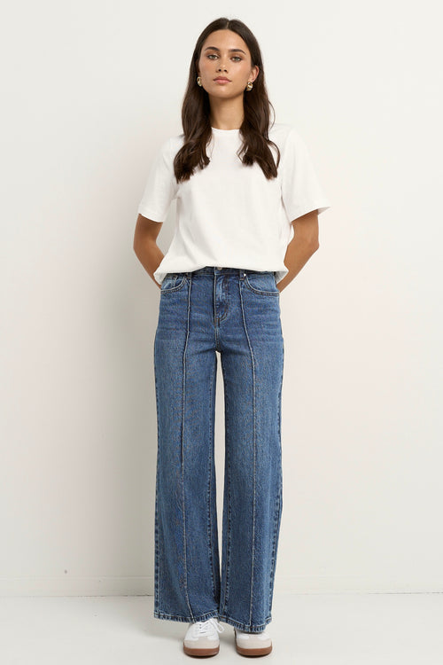 Woman wearing high rise, wide leg classic blue jeans with front seam detail, styled with a white tee and white sneakers.