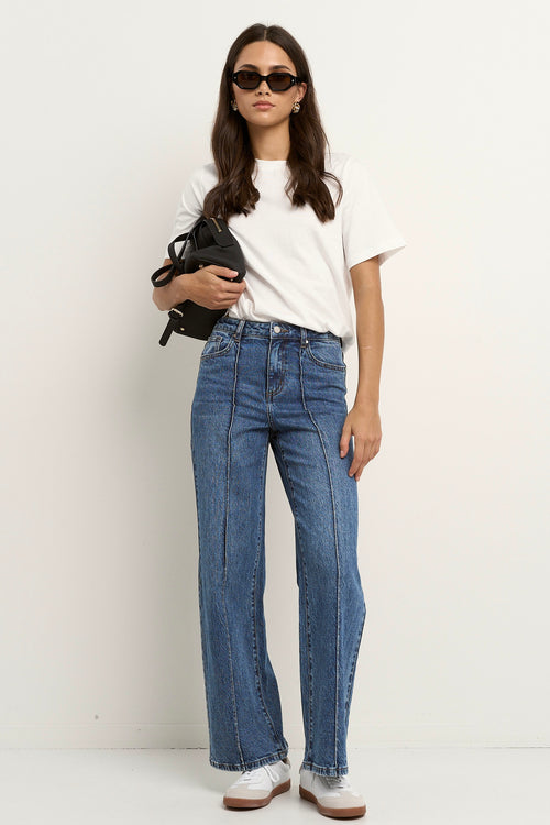 Woman wearing high rise, wide leg classic blue jeans with front seam detail, styled with a white tee and white sneakers.