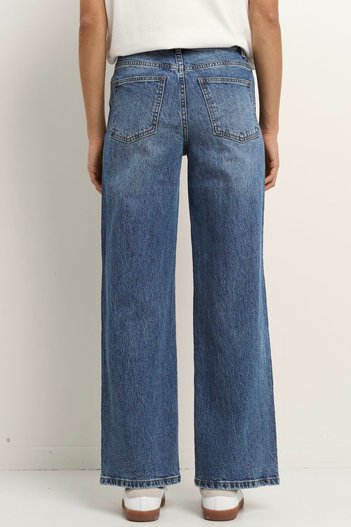 Woman wearing high rise, wide leg classic blue jeans with front seam detail, styled with a white tee and white sneakers.