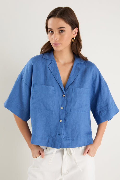 Woman wearing an oversized short-sleeve cornflower blue linen shirt with button front and chest pockets, styled with ecru drawstring jeans.