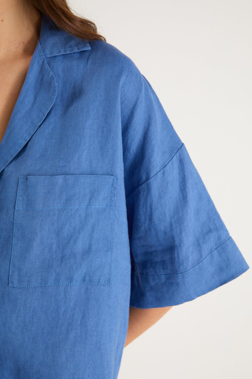 Woman wearing an oversized short-sleeve cornflower blue linen shirt with button front and chest pockets, styled with ecru drawstring jeans.