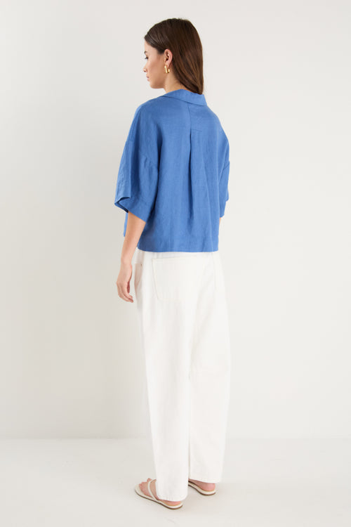 Woman wearing an oversized short-sleeve cornflower blue linen shirt with button front and chest pockets, styled with ecru drawstring jeans.