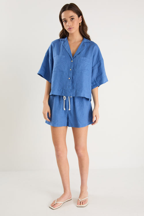 Woman wearing high-rise cornflower blue linen pull-on shorts with rope tie detail, styled with a plain ivory tee.