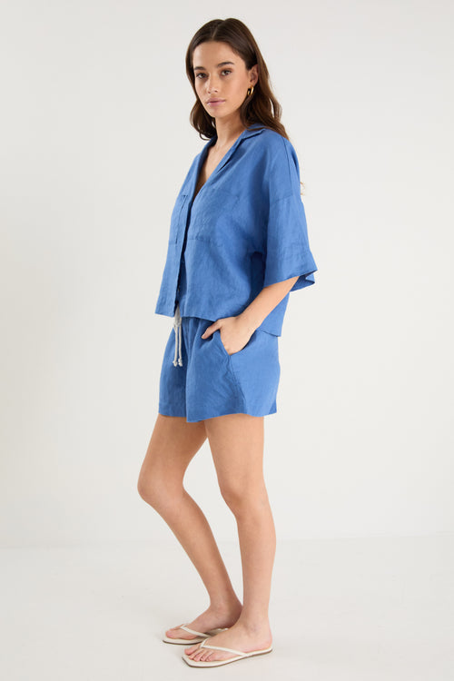 Woman wearing an oversized short-sleeve cornflower blue linen shirt with button front and chest pockets, styled with ecru drawstring jeans.