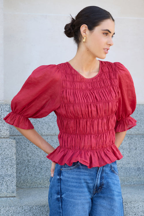 Woman wearing a red blouse with puffed sleeves and blue jeans standing on stone steps.