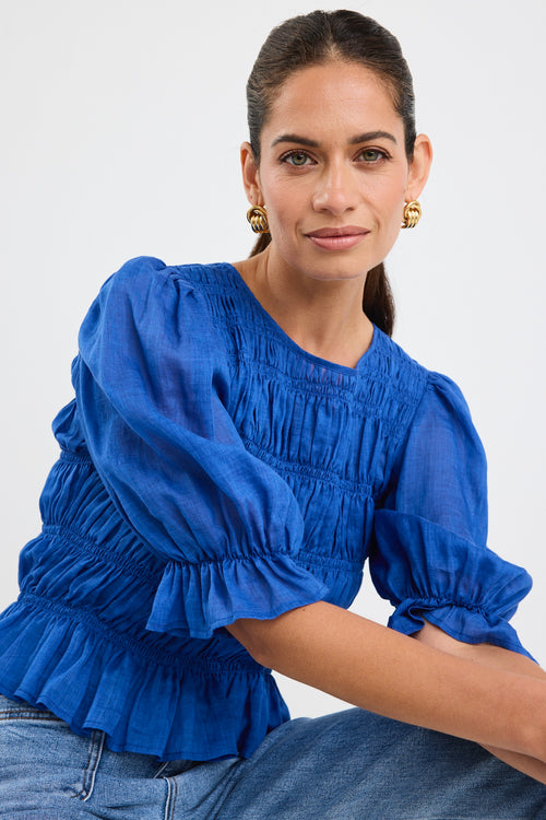 Woman wearing a bright blue ruched cotton top with puff sleeves and peplum hem, styled with light blue jeans.