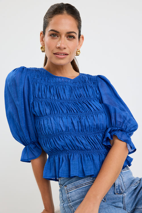 Woman wearing a bright blue ruched cotton top with puff sleeves and peplum hem, styled with light blue jeans.
col_pattern