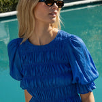 Woman wearing a bright blue ruched cotton top with puff sleeves and peplum hem, styled with white pants.