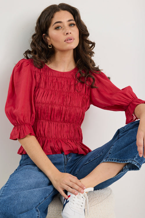 Woman wearing a red blouse and blue jeans sitting on the floor against a plain background