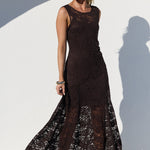 A woman modeling a sleeveless, floor-length dark brown lace dress with a fitted bodice and flared skirt, standing in front of a white arched wall with a strong shadow.