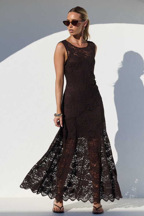 A woman modeling a sleeveless, floor-length dark brown lace dress with a fitted bodice and flared skirt, standing in front of a white arched wall with a strong shadow.