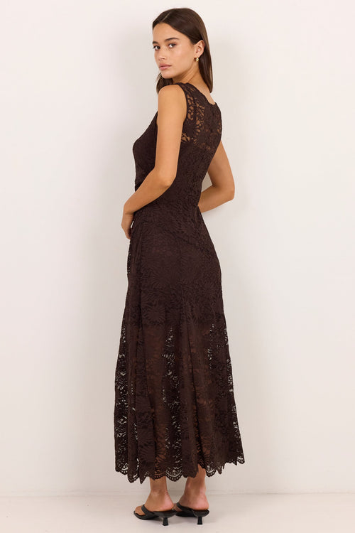 A woman modeling a sleeveless, floor-length dark brown lace dress, shown from a side-back angle against a plain white studio background.