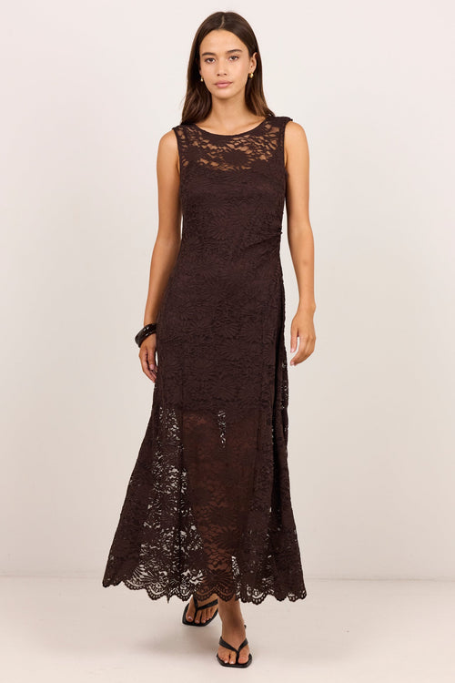 A woman modeling a sleeveless, dark brown lace maxi dress with a high round neckline and a flared skirt, standing against a plain off-white studio background.