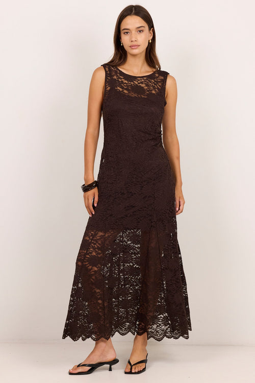 A woman modeling a sleeveless, floor-length dark brown lace dress with a fitted bodice and a flared, textured lace skirt, paired with black flat sandals against a plain off-white studio background.