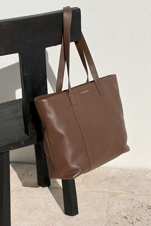 A brown leather tote bag with long shoulder straps, hanging from the backrest of a black wooden chair against a textured off-white wall and light stone floor.