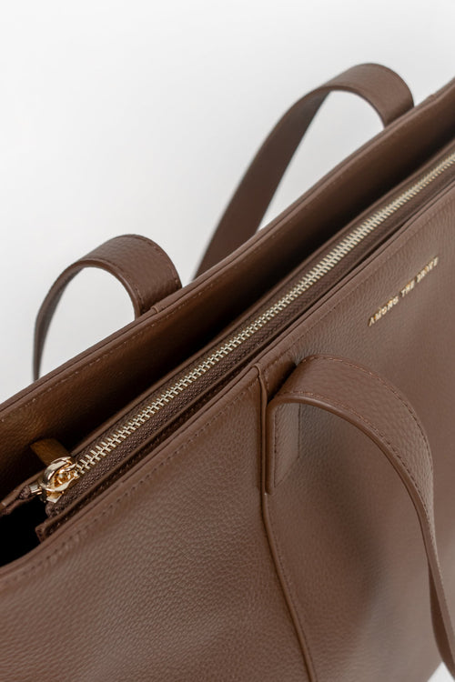 A close-up of a brown pebbled leather tote bag with gold-tone hardware, showing the interior zipper and a small gold brand logo on the side.
