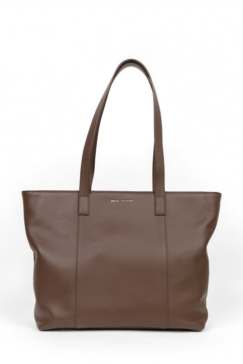 A brown leather tote bag with long shoulder straps and a small gold logo plaque on the front, photographed against a plain white background.