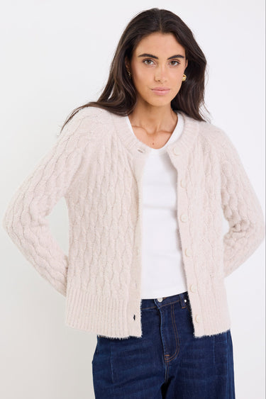 Woman wearing a beige cable knit cardigan over a white shirt with blue jeans on a white background