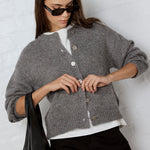 Woman wearing a gray cardigan over a white shirt, holding a black bag against a light background