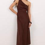 A woman modeling a dark brown one-shoulder midi dress with a draped neckline, standing against a plain white studio background.
