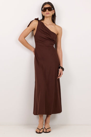 A woman modeling a dark brown one-shoulder midi dress with a draped neckline, standing against a plain white studio background.