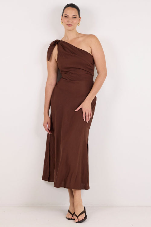 A woman modeling a dark brown one-shoulder midi dress with a draped, gathered bodice and a flowing skirt, standing against a plain white studio background.