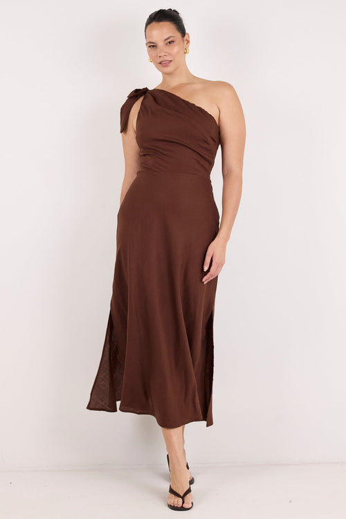 A woman modeling a brown one-shoulder midi dress with a draped, gathered bodice and a flowing skirt, paired with black strappy heels, against a plain white studio background.