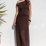 A woman wearing a dark brown off-the-shoulder maxi dress and sunglasses stands against a white stucco wall next to a potted plant.