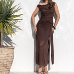 A woman wearing a sleeveless brown maxi dress and sunglasses stands next to a white wall beside a pool, with a potted plant and trees in the background.