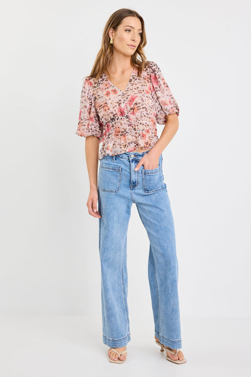 Woman wearing a short sleeve bubble blouse with a mix of floral and animal prints, button front, and elastic waist, styled with light blue jeans.