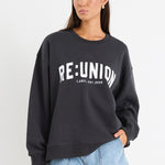 Woman wearing a charcoal grey oversized sweatshirt with "RE:UNION" printed across the chest, styled with light blue denim shorts.