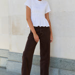 Woman wearing a white top and brown pants standing against a light-colored wall.