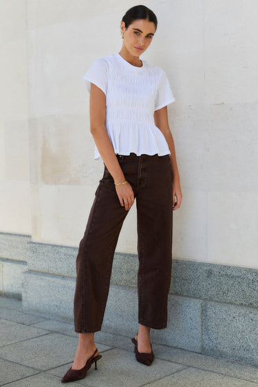 Woman wearing a white top and brown pants standing against a light-colored wall.