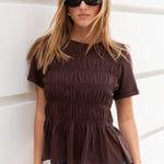 Woman wearing a brown peplum top and sunglasses against a white wall.