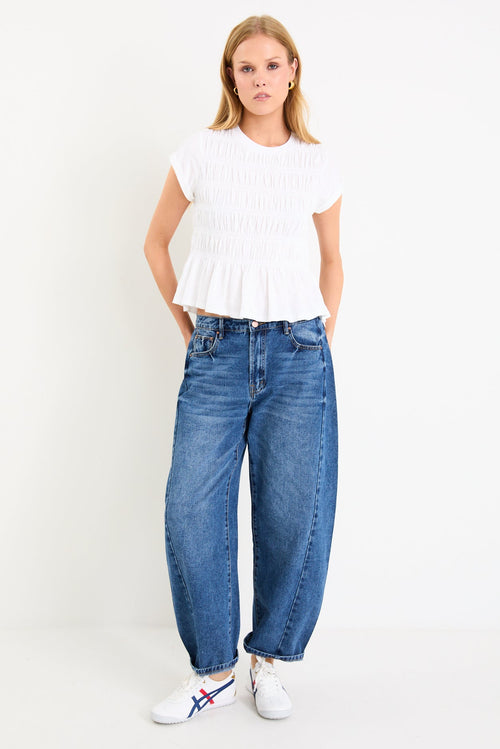Model wearing a white short sleeve shirred cotton top with a peplum hem and crew neckline