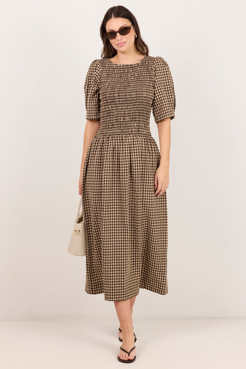 A woman modeling a brown and beige checkered midi dress with puff sleeves and a smocked bodice, standing against a plain light gray studio background.
